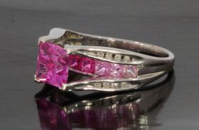 Moderne witgouden dames ring met een princess geslepen synthetische roze saffier van 7mm waaronder een rij van vierkante gefacceteerde in kleur oplopende lichtroze tot dieproze saffieren. De chaton zetting van de middensteen gedecoreerd met in totaal 16 b